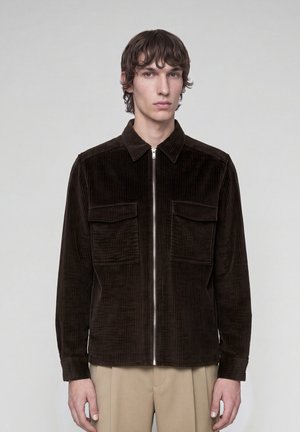 Young man wearing a dark brown corduroy jacket with front pockets and a zipper, paired with beige trousers, standing against a plain background.