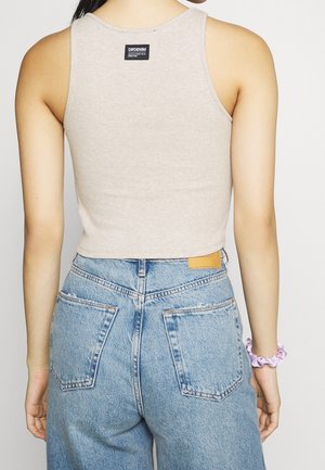 Kvinde iført en beige crop-top med sort mærke og lyseblå højttaljede jeans, stående med armene afslappet ned langs siden.