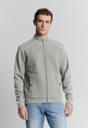 Young man with short curly hair wearing a light gray textured zip-up jacket and blue jeans, standing against a plain light background.