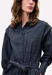 Robe en denim sombre avec des rayures verticales bleues, un col, un bouton devant et une poche poitrine. Elle a des manches longues et une ceinture en tissu.
