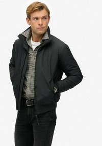 Chaqueta de abrigo negra con un alto cuello, cierre de cremallera y puños acanalados, superpuesta a una camisa de cuadros, llevada con jeans negros.