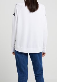 Vit sweatshirt med lös passform, raglanärmar, ribbade ärmslut och rak nederkant, i kombination med blå jeans. Slät textur.