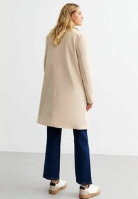 Beige oversized jas met een rechte snit, gemaakt van gladde stof. Gecombineerd met donkerblauwe wijde jeans en witte sneakers.