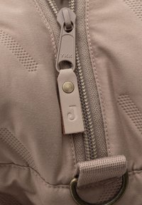 Beige stof close-up met een YKK-rits en een leren lus met het gestempelde letter "J," en een metalen ringaccent. Gelaagd patroon op de stof.