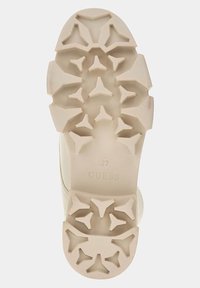 Beige rubber sole with textured surface, featuring a geometric pattern of prominent shapes for traction. Brand logo "GUESS" embossed.