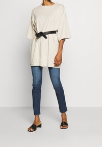 Beige oversize t-paita lyhyillä hihoilla, musta nahkavyö, yhdistetty tummansinisiin istuviin farkkuihin ja mustiin liukuvaan kenkään.