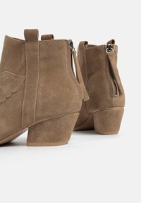 Bottines en daim marron avec un petit talon, dotées de fermetures éclair latérales et de coutures décoratives sur l'extérieur. Texture douce et forme arrondie.