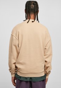 Felpa beige oversize con spalle cadute, polsini a costine e una texture morbida, indossata sopra una camicia verde, abbinata a pantaloni viola larghi.