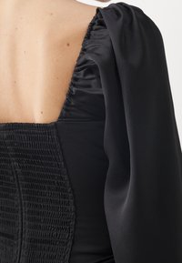 Abito nero con schiena arricciata e maniche a sbuffo, che mette in evidenza la morbida trama del satin e una silhouette aderente.