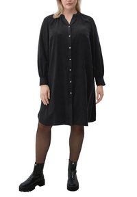 Femme portant une robe noire à boutons, longueur genou, à manches longues, des collants noirs transparents et des bottines à plateforme noires avec des zips.