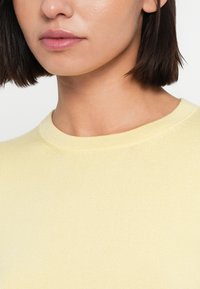 Κίτρινο ribbed πουλόβερ με στρογγυλή λαιμόκοψη, με ομαλή υφή και εφαρμοστή σχεδίαση. Κοντινή λήψη που εστιάζει στη λαιμόκοψη και τις λεπτομέρειες του υφάσματος.