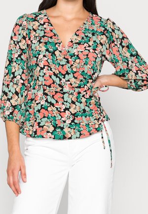 Vrouw draagt een zwarte wikkelblouse met koraal-, groene en beige bloemenprint en driekwart pofmouwen, gecombineerd met witte broek.