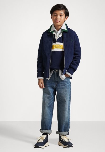 Jaka ar quiltētiem rakstiem tumši zilā krāsā, virs svītrainā tumši zila un dzeltena polo krekla, ar gaiši zilu krekla šuvīti zem tā. Izbalējuši zili džinsi un tumši zilas kedas noslēdz apģērbu.