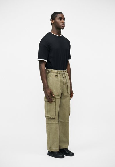 Άνδρας που στέκεται στο πλάι φορώντας μαύρο T-shirt με άσπρη ένωση, χακί παντελόνι cargo με μεγάλες τσέπες και μαύρα παπούτσια σε λευκό φόντο.