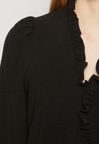 Black textured blouse featuring a ruffled neckline and puffed sleeves, with a smooth finish and delicate stitching details.