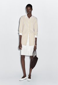 White and yellow striped shirt with long sleeves, paired with a shiny white skirt. Holding a dark brown bag. Footwear is white.