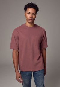 T-shirt a maniche corte color bordeaux in cotone, con taschino frontale, scollatura rotonda e vestibilità rilassata, abbinato a jeans blu.