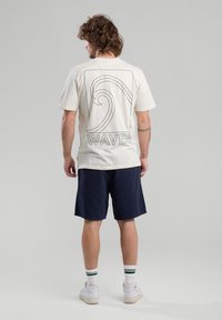 T-shirt en coton crème avec un design de vagues noires et le texte "WAVES" au dos, porté avec un short bleu marine et des baskets blanches avec des rayures vertes.
