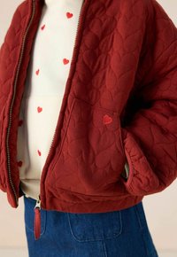 Rode gewatteerde jas met een ritssluiting, met hartvormige patronen. Zak versierd met een klein hartdetail. Draagt over een crèmekleurig shirt met hartaccenten.