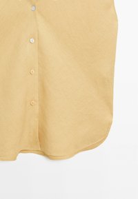 Camicia gialla chiaro, con bottoni e una texture morbida, con orlo arrotondato. Presenta diversi bottoni rivestiti in tessuto lungo la parte frontale.