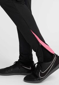 Pantalones deportivos negros con una cremallera rosa a lo largo del costado. Combinados con zapatillas deportivas negras con detalles en gris y acabado texturizado.