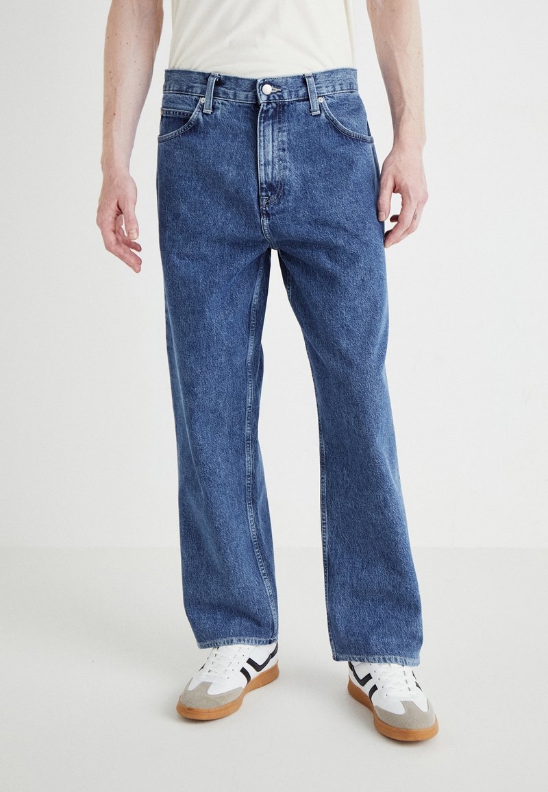 Mann trägt lockere blaue Jeans und weiße Sneakers mit schwarzen Streifen, steht vor einem einfarbigen weißen Hintergrund.