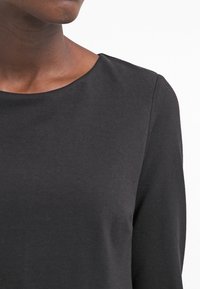 Svart långärmad topp med rund halsringning, tillverkad av mjukt tyg. Har en slät struktur och en figursydd siluett. Inga ytterligare mönster.