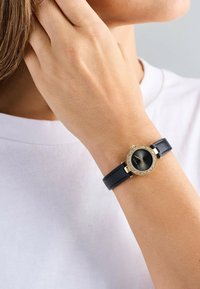 Round watch with a black leather strap, gold-tone case, and black dial, featuring clear crystal accents around the bezel.