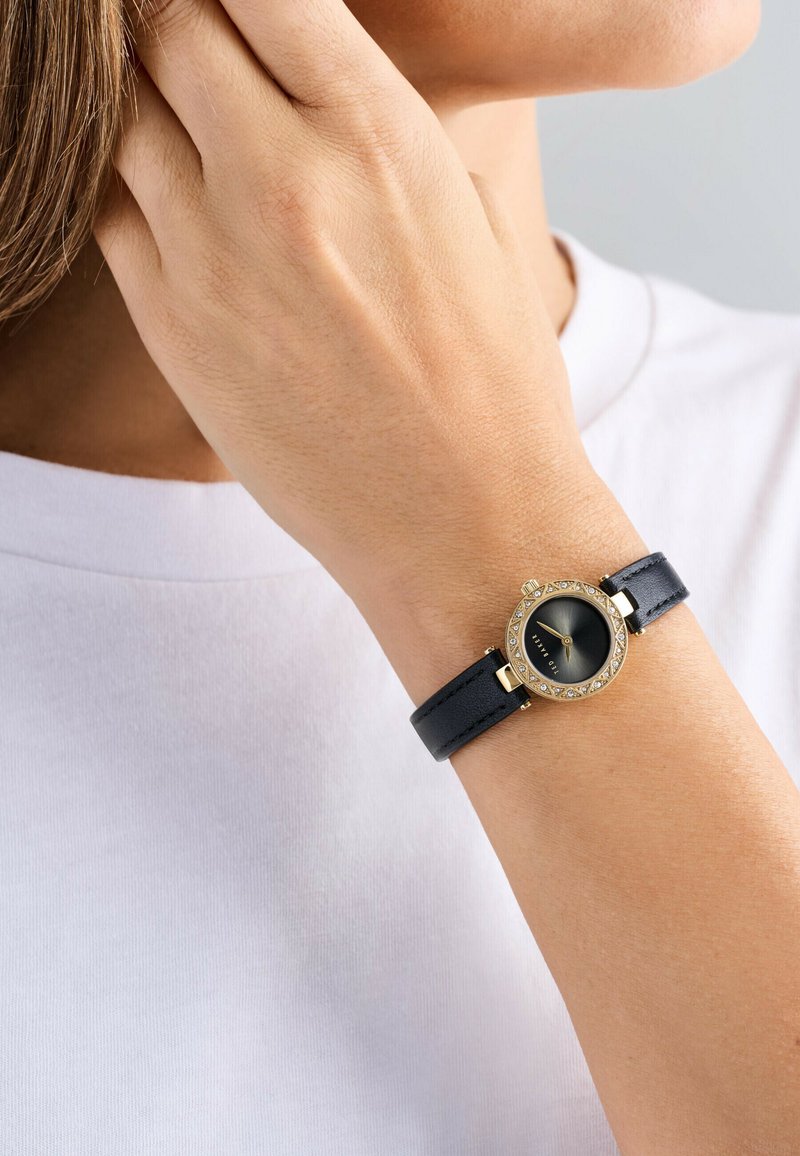 Runde Uhr mit einem schwarzen Lederarmband, Gehäuse in Goldfarbe und schwarzem Ziffernblatt, das klare Kristallakzente um die Lünette bietet.