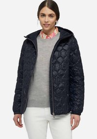 Veste matelassée navy avec zip à l'avant, poignets élastiques et capuche. Portée par-dessus un pull gris clair, avec une chemise colorée à col en dessous.