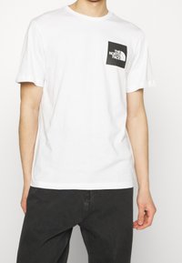 Man med en vit kortärmad T-shirt med en svart fyrkantig North Face-logga på vänster bröst, i kombination med svarta byxor.