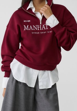 Persoon draagt een bordeauxrode kwartritssweatshirt met de tekst "Manhattan Upper East Side New York" over een wit overhemd met knopen en grijze broek.