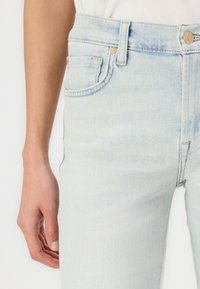 Jean taille haute bleu clair en denim avec un léger délavage, cinq poches et des accents en métal doré au niveau de la taille.