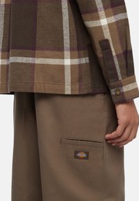 Una camicia oversize a quadri marroni in morbido tessuto, con strisce bianche e viola. Abbinata a pantaloni beige, che mostrano una tasca con logo.