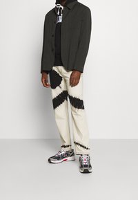 Giacca grigio scuro, felpa nera con logo, pantaloni bianchi con motivi tie-dye neri e scarpe da ginnastica grigie con dettagli rossi.