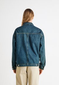 Denimjack in donkerblauw met een rechte snit, knoopmanchetten en twee zijzakken. De achterkant heeft een centrale naad en geen versieringen.