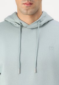 Hoodie em azul-teal claro feito de material macio, apresentando um capuz com cordão e um logo em relevo na parte frontal. Textura suave com detalhes em prateado.