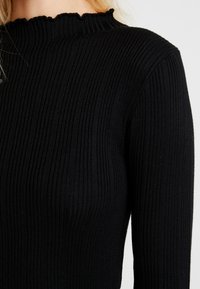Schwarzes geripptes Langarmoberteil mit hohem Rüschenausschnitt. Der Stoff hat eine weiche Textur mit vertikalen Linien, die sich durchgehend ziehen.