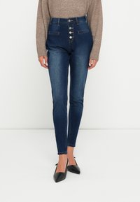 Jeans ajustados de talle alto en denim azul oscuro, con cierre frontal de cinco botones, costuras en contraste y dos bolsillos delanteros.