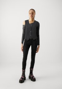 Chaleco negro con dobladillo en pico y tres botones, llevado sobre una blusa de manga larga oscura y combinado con pantalones negros texturizados y botas negras gruesas.