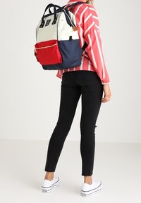 Wielokolorowy plecak z białymi, czerwonymi i granatowymi sekcjami, wykonany z teksturowanej tkaniny, z podwójnymi uchwytami na górze oraz wyraźnym akcentem zamka błyskawicznego.