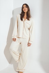 Kremkrāsas pidžamu komplekts, kas izgatavots no mīkstas auduma; tam ir garas piedurknes, pogājams top un platās bikses ar kontrastējošu apdari.