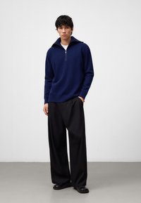 Pullover lavorato a maglia blu navy con colletto alto e chiusura a mezza zip, abbinato a pantaloni neri larghi e scarpe nere slip-on. Texture semplice.