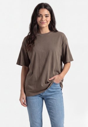 Ung kvinde med langt, mørkt hår iført en løs brun t-shirt og lyseblå jeans, står med den ene hånd i lommen.
