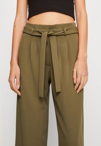 Oliwkowe spodnie z wysokim stanem, charakteryzujące się szerokimi nogawkami, plisami oraz pasującym paskiem z materiału związanym z przodu. Gładki, lekki materiał.