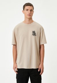 Camiseta de manga corta color beige, hecha de una tela suave. Presenta un logo gráfico negro en el lado izquierdo del pecho. Corte relajado y escote redondo.