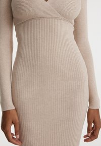 Abito a maglia a maniche lunghe in una tonalità beige chiaro. Presenta una texture a coste e un collo avvolgente, enfatizzando una silhouette aderente.