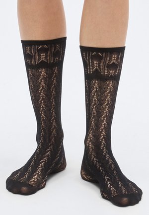Chaussettes en dentelle noire avec un motif floral, design jusqu'aux genoux et finition texturée avec des sections transparentes pour un intérêt visuel renforcé.