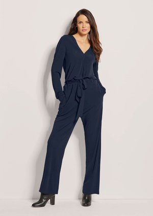 Frau steht mit den Händen in den Taschen, trägt eine marineblaue Langarm-V-Neck-Bluse und passende weite Hosen mit schwarzen Stiefeletten.