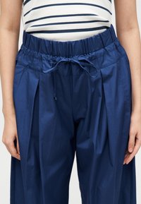 Personne portant un pantalon taille haute bleu marine avec une taille élastique à cordon et un haut rayé blanc et bleu marine rentré dedans.
